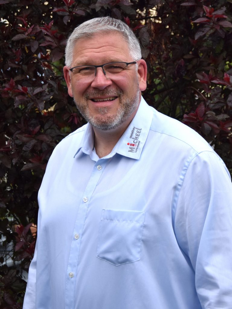 Portrait von dem Geschäftsführer Thorsten Micheel. Er trägt ein weißes Hemd das mit dem Firmenlogo bestickt ist, hat graue Haare, eine schwarze Brille und einen Bart. Er steht vor einer Hecke und lächelt in die Kamera.