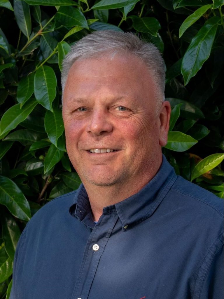 Portrait von dem Geschäftsführer Holger Micheel. Er trägt ein dunkelblaues Hemd und hat graue Haare. Er steht vor einer Hecke und lächelt ebenfalls in die Kamera.